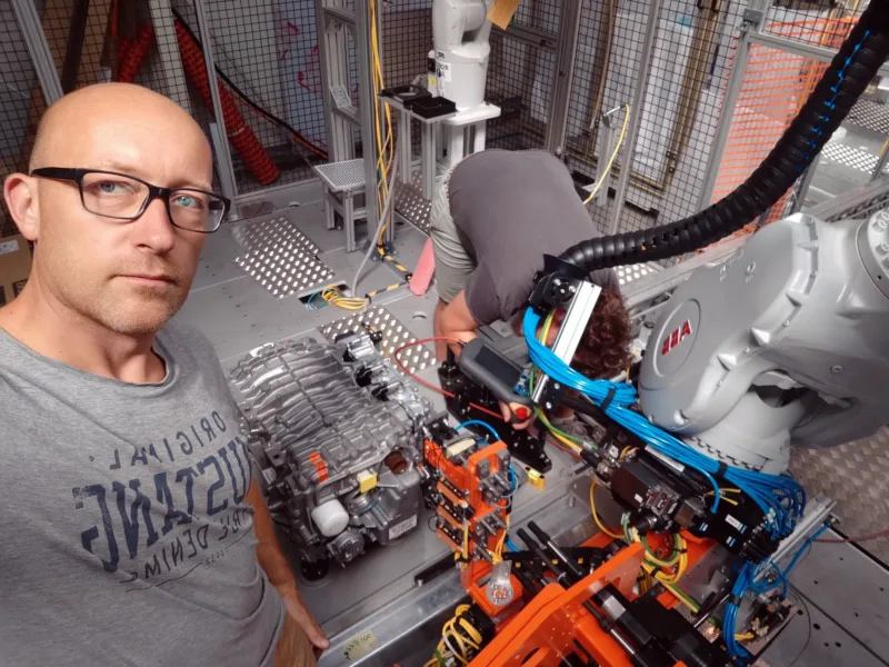 Mężczyzna w okularach robi sobie selfie w laboratorium, w którym znajdują się roboty przemysłowe i maszyny firmy Konstrukcje Mechaniczne. Inna osoba pochyla się nad sprzętem, pracując przy nim. Teren jest ogrodzony metalowym płotem ze względów bezpieczeństwa. Mężczyzna w okularach robi sobie selfie w laboratorium, w którym znajdują się roboty przemysłowe i maszyny firmy Konstrukcje Mechaniczne. Inna osoba pochyla się nad sprzętem, pracując przy nim. Teren jest ogrodzony metalowym płotem ze względów bezpieczeństwa.