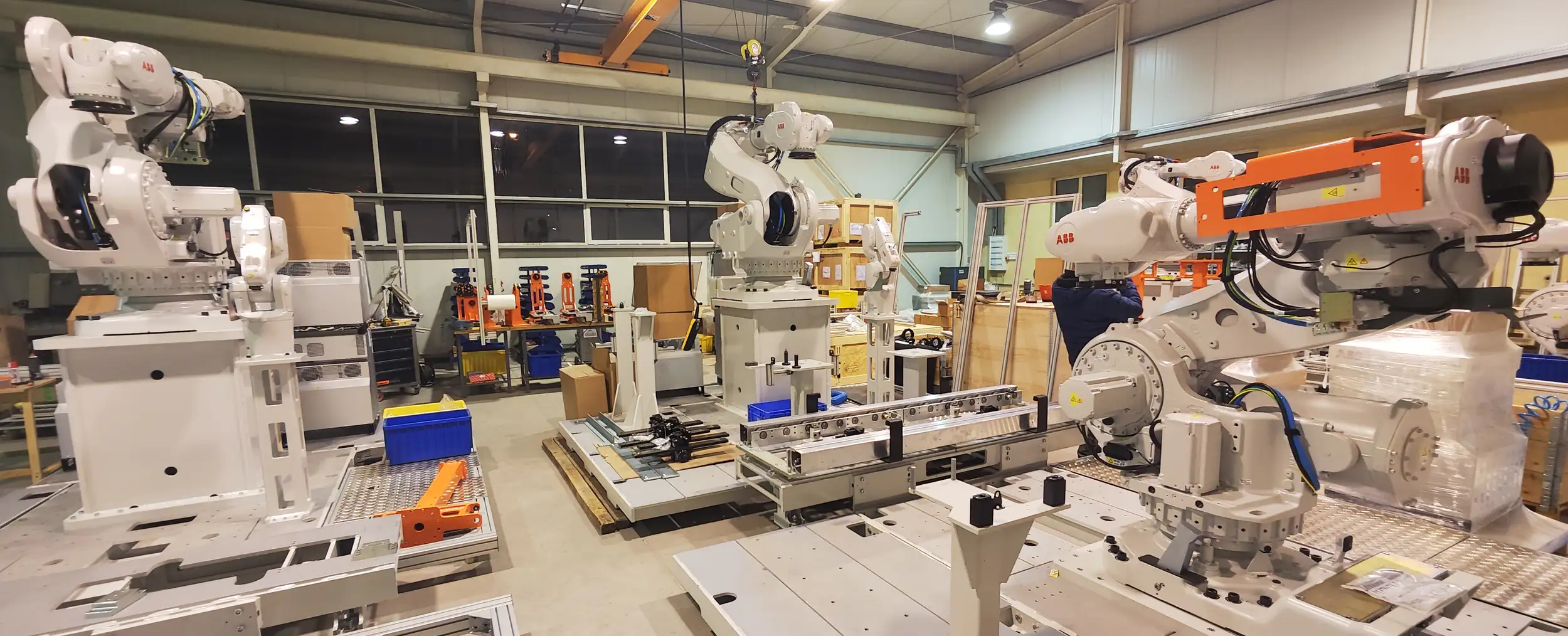 Roboty przemysłowe montują maszyny w dobrze oświetlonej przestrzeni produkcyjnej, w której widać różne urządzenia, pudełka i skrzynie magazynowe. W tle widać wysokie sufity i duże okna.